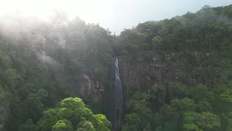 Moving-through-a-sun-drenched-misty-fog-revealing-a-hidden-tropical-rainforest-waterfall