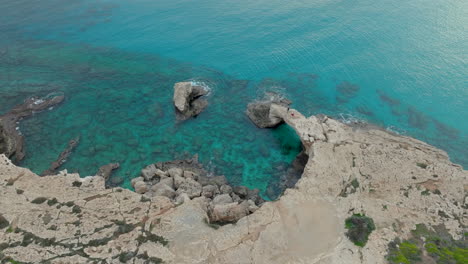 Aérea:-La-Formación-Rocosa-Natural-Conocida-Como-El-Puente-Del-Amor,-Ubicada-En-Ayia-Napa,-Chipre,-Aguas-Cristalinas-De-Color-Turquesa-Rodean-El-Arco-De-Piedra-Caliza-Erosionada,-Sorprendente-Contraste-Con-La-Escarpada-Costa