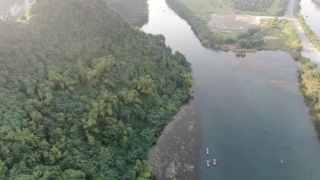 Drone-aerial-view-in-Vietnam-flying-over-a-valley-surrounded-by-rocky-mountains-covered-with-green-trees-over-a-river-with-small-boats-in-Ninh-Binh-on-a-sunny-day