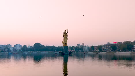 Maravíllate-Ante-La-Majestuosa-Vista-Frontal-De-La-Inmensa-Estatua-Dorada-Del-Señor-Shiva-En-El-Lago-Sursagar,-Vadodara,-Mientras-Cae-El-Anochecer,-Ofreciendo-Un-Impresionante-Espectáculo-De-Grandeza-Divina.