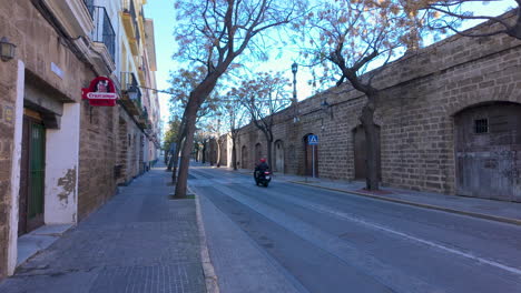 A-view-from-a-TukTuk-shows-a-quiet-street-in-Cádiz,-with-a-motorcyclist-riding-past-traditional-stone-buildings-and-a-clear-blue-sky-above