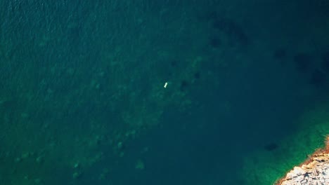 Vista-De-Pájaro-Del-Barco-Flotando-En-Medio-Del-Agua-Azul-Profunda-Del-Océano-Con-Una-Superficie-Rocosa-Debajo