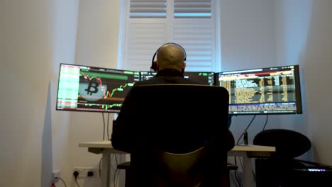 Bald-man-analyzing-stock-market-trends-on-multiple-monitors-in-a-dark-room,-backlit-by-screens