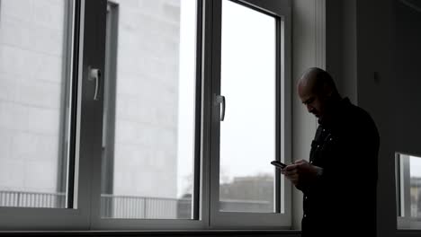 A-man-stands-by-the-window-with-gloomy-weather-outside,-engrossed-in-typing-on-his-mobile-phone-screen