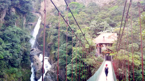Person-walking-on-suspension-bridge-in-Baños,-Ecuador-with-a-waterfall-and-lush-greenery-in-the-background,-serene-and-adventurous