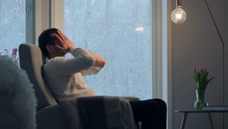 Mann-Im-Pullover-Fühlt-Sich-Gestresst-In-Einem-Gemütlichen-Zimmer,-Während-Draußen-Schnee-Fällt,-Hand-Auf-Dem-Kopf,-Nachdenkliche-Stimmung,-Stimmungsvolles-Licht