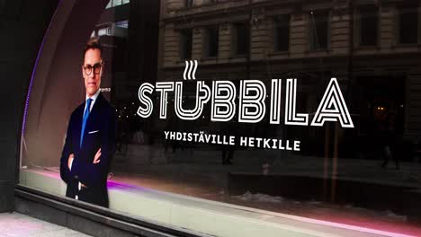 President-of-Finland-Alexander-Stubb-election-office-in-Helsinki-downtown,-man-in-suit-by-window-with-'STUBBILLA'-neon-sign,-evening-city-backdrop,-Helsinki-vibe