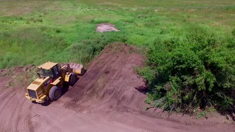 A-bulldozer-at-work-in-a-green-field,-removing-grass-on-a-sunny-day,-aerial-view