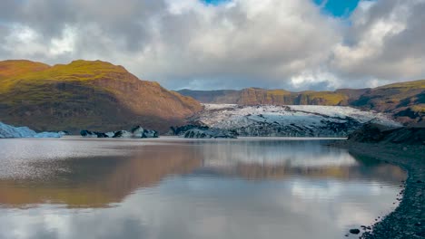Sunlit-Sólheimajökull-glacier-in-Iceland-reflecting-in-a-serene-lake,-with-a-dramatic-sky