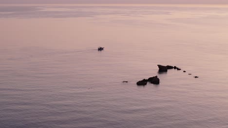 Fischerboot-Navigiert-Zwischen-Freiliegenden-Felsen-Ins-Offene-Meer,-Während-Das-Wasser-Orange-Glitzert