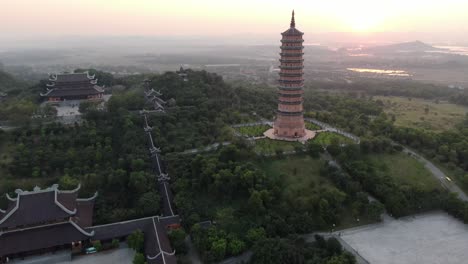 Drone-aerial-view-in-Vietnam-flying-over-a-buddhist-temple-area-and-pagoda-filled-with-green-trees-in-front-of-the-sun-in-Ninh-Binh-at-sunset