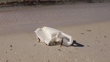 Weathered-plastic-bottle-littered-on-sandy-beach-with-waves-in-background