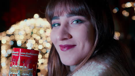 Close-up-of-a-beautiful-Woman-with-dark-Hair-saying-cheers-with-her-hand-and-drinking-Punch-from-red-Heart-Cup-at-Vienna-Christmas-Market-,-surrounded-by-Christmas-Lights-in-the-background
