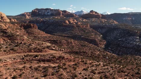 A-high-flying-drone-shot-over-a-remote-dirt-road-cutting-through-the-vast-and-unique-desert-land-near-Moab,-Utah,-with-the-snowy-Rocky-Mountains-towering-in-the-distance