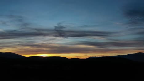 drone-flight-in-a-valley-visualizing-the-sunset-with-all-the-dark-mountains-a-blue-yellow-and-orange-sky-with-clouds-with-a-tilt-turn-of-the-camera-upwards-in-Avila-Spain