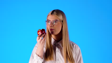 Attractive-young-woman-takes-a-bite-of-an-red-apple,-blue-studio-background