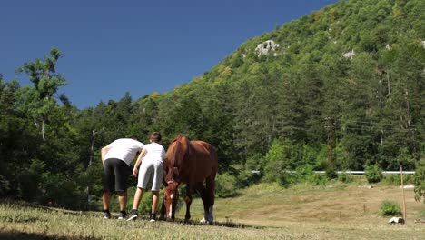 Father-and-son-petting-chestnut-horse-on-field-in-hilly-Balkan-landscape