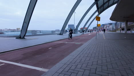 Pedestrians-and-cyclists-navigate-a-wide-pathway-under-the-modern-steel-arches-near-Amsterdam's-waterfront,-showcasing-the-blend-of-architecture-and-active-transit