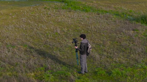 Male-Photographer-Carrying-Camera-On-Tripod-On-The-Field-Near-Lake