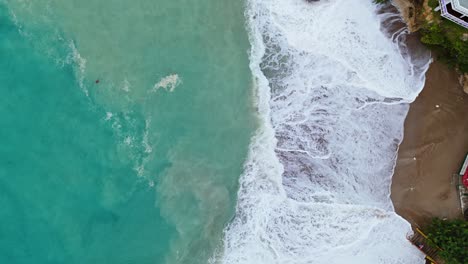 Dark-brown-sand-in-cove-churns-in-undertow-of-strong-wave-swell-in-ocean,-drone-bird's-eye-view