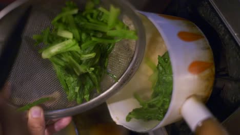 Freshly-cooked-green-vegetables-moved-from-metal-strainer-into-small-white-cooking-pot,-filmed-as-vertical-slow-motion-closeup-shot-in-handheld-style