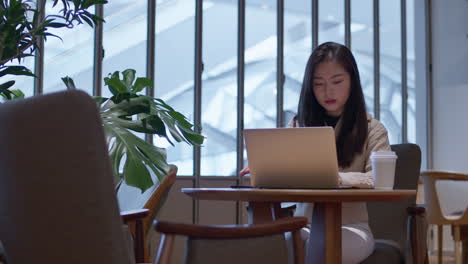Hübsches-Asiatisches-Mädchen-Arbeitet-Im-Café-Am-Laptop-Und-Checkt-Nachrichten-Auf-Ihrem-Telefon