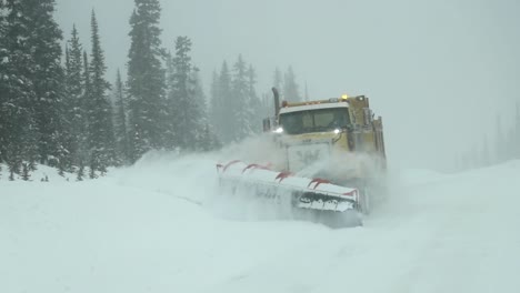 Yellow-Canadian-snow-plow-truck-driving-towards-the-camera-while-clearing-the-countryside-road-off-freshly-fallen-snow-on-a-snowy-overcast-day-in-slow-motion