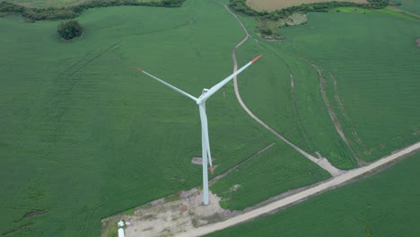 Aerial-shot-of-a-solitary-wind-turbine-in-lush-green-field,-dirt-access-road-visible,-overcast-day