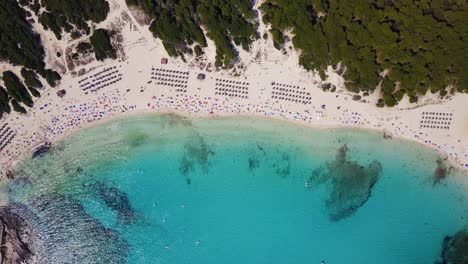 Cala-agulla,-vibrant-beach-with-tourists,-clear-turquoise-water,-and-umbrellas,-aerial-view