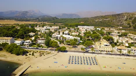 Drone-panning-from-the-right-to-the-left-side-of-the-frame-showing-the-expanse-of-Playa-del-Port-de-Pollença's-beachfront,-a-tourist-destination-in-Mallorca,-Spain