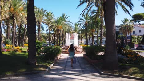 Young-woman-walks-in-park-with-fountain-in-summertime-Antibes,-tilt-up