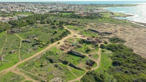 Aerial-view-sprawling-archaeological-site-of-the-Tombs-of-the-Kings-in-Paphos,-Cyprus,-nestled-between-the-cityscape-and-the-coast,-highlighting-the-ancient-ruins-amid-modern-urban-development