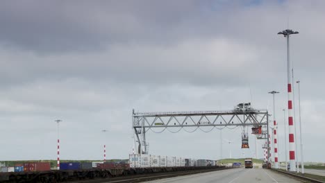 Freight-train-passing-under-a-gantry-crane-at-a-container-terminal,-overcast-sky