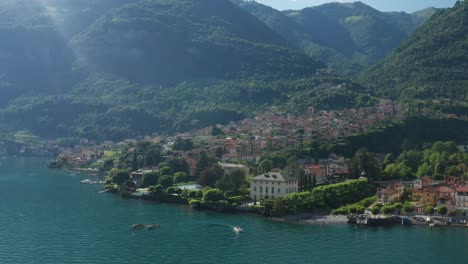 Una-Exuberante-Vegetación-Rodea-El-Lago-Como-En-Ossuccio,-Italia,-Los-Barcos-Se-Desplazan-Suavemente,-El-Telón-De-Fondo-De-La-Montaña