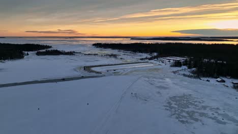 A-snow-covered-harbor-at-sunset-with-golden-skies-and-sparse-trees,-aerial-view
