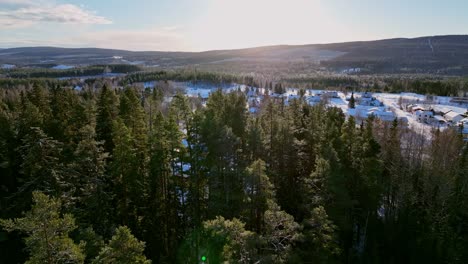 Winter-forest-aerial-view-with-a-glimpse-of-Skorped,-Sweden-village,-sunlight-piercing-through-trees