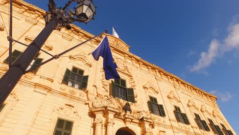 Low-angle-shot-of-the-Castille-palace-and-the-street-lights-with-two-flags,-Maltese-and-European-union,-fluttering-on-the-wind