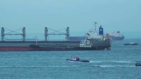 Small-government-authority-boats-sailing-around-the-large-cargo-ships-on-the-strait-of-Singapore,-inspecting-the-custom-goods-and-documenting-it-before-it-arrives-to-inland,-close-up-shot