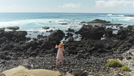 Mujer-Caminando-Sobre-Rocas-Negras-Cerca-De-La-Orilla-Del-Mar-Mientras-Usa-Un-Vestido-Floral,-Cámara-Lenta-Estática