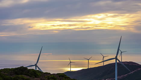 Windturbinen-Drehen-Sich-Im-Wind-Und-Erzeugen-Strom-Vor-Einem-Sonnenuntergang-Mit-Orangefarbenem-Himmel-Und-Ziehenden-Wolken-über-Der-Landschaft-Und-Dem-Meer.