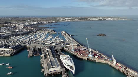 St-Peter-Port-Guernsey-flight-over-ferry-terminal-with-ferry-in-dock,-commercial-dock-with-cranes-towards-Salerie-Corner-and-over-QE-II-Marina-with-views-over-Belle-Greve-Bay-on-bright-sunny-day