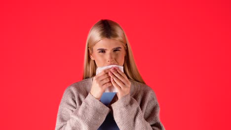 Caucasian-young-blonde-woman-sneezes-into-a-tissue-with-a-cold,-portrait-shot-with-endless-red-studio-background