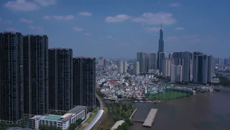 Ho-Chi-Minh-City,-Saigon-River-and-Binh-Thanh-skyline-on-sunny,-clear-day-featuring-landmark-building-and-golden-river-from-drone-tracking-shot