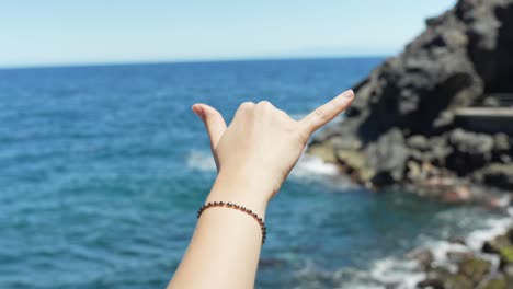 Person-hand-showing-cool-gesture-near-rocky-ocean-coastline,-POV