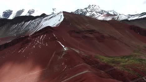 Drone-view-in-Peru-flying-over-red-valley-in-Cuzco,-showing-red-colored-mountains-surrounded-by-snowed-mountains-in-the-horizon