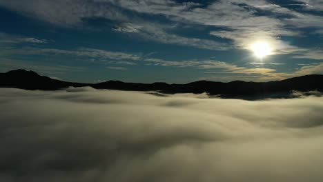 Drohnenflug-über-Einem-Wolkenmeer-Mit-Der-Sonne-Im-Vordergrund,-Der-Die-Berge-Visualisiert,-Die-Im-Licht-Dunkel-Aussehen,-In-Einer-Rechtskurve-Mit-Blauem-Himmel-Bei-Einem-Sonnenaufgang-Im-Winter-In-Avila,-Spanien