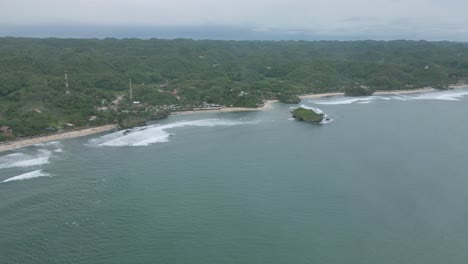 A-lush-indonesian-beach-with-waves-and-a-solitary-islet,-cloudy-day,-aerial-view