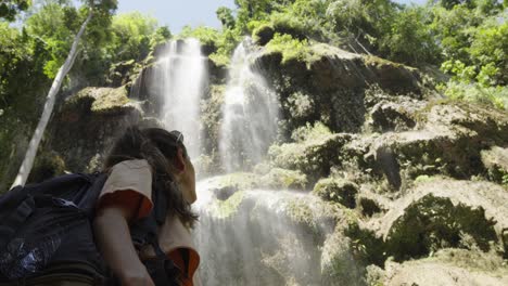 Young-caucasian-woman-with-adventuring-backpack-stares-up-at-tropical-Tumalog-waterfall-in-Phillippines