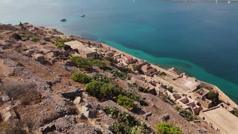 Drone-push-in-descends-over-cliffside-along-Spinalonga-historic-ruins-into-sweeping-view-of-blue-bay