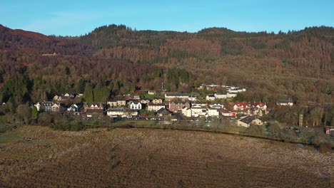 Panorámica-Aérea-Panorámica-A-Través-De-Los-Campos-Con-Vistas-Al-Pueblo-De-Aberfoyle-Y-Al-Fondo-De-La-Ladera-En-Escocia-Durante-El-Otoño-En-Un-Día-Soleado